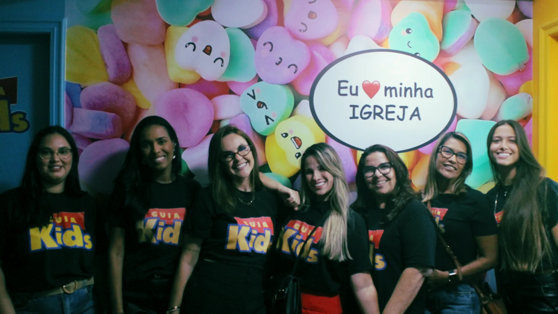 A equipe do GUIA Kids reunida, de camiseta preta, diante do painel "Eu amo minha igreja"
